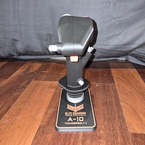 May include: Black joystick with red and silver accents. The base is black with gold lettering that reads "ELITE SQUADRON MASTERBUILDS A-10 THUNDERBOLT II". The joystick is designed for flight simulation games.