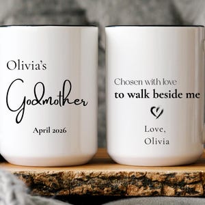 May include: Two white ceramic mugs with black handles and rims. The first mug features the text Olivia's Godmother and April 2026. The second mug displays the text Chosen with love to walk beside me, a heart icon, and Love, Olivia.
