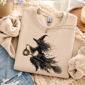 May include: A beige crewneck sweatshirt featuring a detailed black ink-style illustration of a witch flying on a broomstick while holding a glowing lantern. The garment has a ribbed collar and a Gildan brand tag visible at the inner neckline.