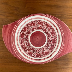 Puede incluir: Un plato de servir grande, de forma ovalada, con un borde blanco y un centro rojo. El centro presenta un diseño floral blanco.