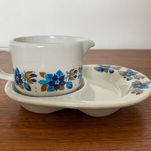 May include: A white ceramic creamer with a blue and gold floral pattern sits on a matching saucer with a scalloped edge.
