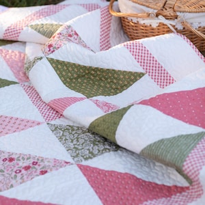 May include: A folded quilt with a patchwork design featuring white, pink, and green triangles. The quilt has a textured surface and is decorated with floral and gingham patterns. A wicker picnic basket is in the background.