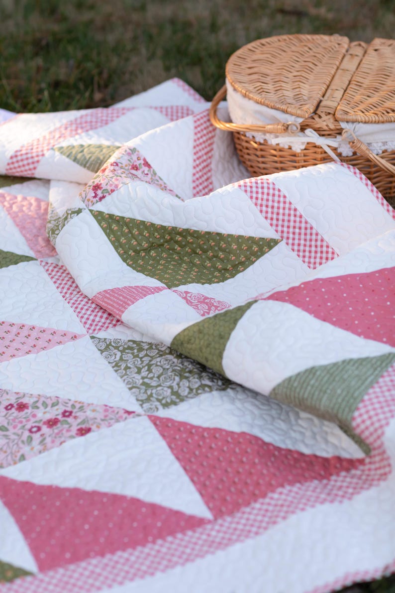 May include: A folded quilt with a patchwork design featuring white, pink, and green triangles. The quilt has a textured surface and is decorated with floral and gingham patterns. A wicker picnic basket is in the background.