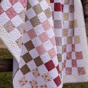 May include: Handmade quilts with a checkerboard design. The quilts display squares of white, pink, brown, and floral fabrics. The quilts are draped over a wooden fence, highlighting the detailed quilting. The quilts are made with high-quality materials.
