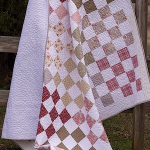 Op de afbeelding: Een handgemaakte quilt met een witte, gewatteerde achterkant en een patchwork-ontwerp. Het patchwork heeft afwisselende vierkanten van witte, bordeauxrode, roze en bruine stoffen. De quilt is over een houten hek gedrapeerd.