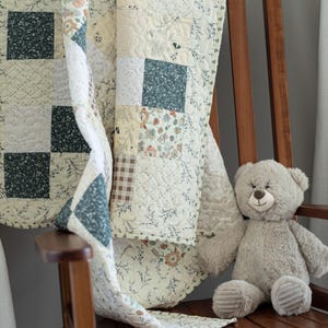 May include: A patchwork quilt featuring a variety of fabric squares in shades of cream, dark green, and brown, with floral, plaid, and solid patterns. The quilt has a quilted texture and is draped over a wooden chair next to a plush beige teddy bear.