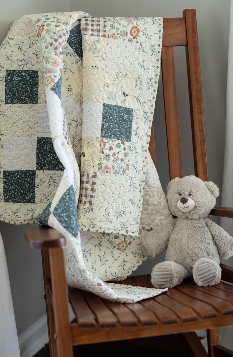 May include: A patchwork quilt featuring a variety of fabric squares in shades of cream, dark green, and brown, with floral, plaid, and solid patterns. The quilt has a quilted texture and is draped over a wooden chair next to a plush beige teddy bear.