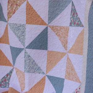 May include: A quilted blanket with a geometric design. The quilt features white, peach, and blue triangles, some with floral patterns. The edges are finished with a blue border.
