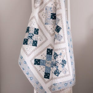 May include: A white and blue floral quilt draped over a white ladder. The quilt features a patchwork design with squares of blue floral patterns and white fabric. The edges are trimmed with a blue floral border, creating a cozy, handcrafted aesthetic.