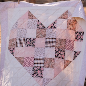 May include: A handmade quilt featuring a heart design. The heart is composed of various fabric patterns in shades of pink, brown, and black, set against a white background. The quilt is bordered with a floral pattern and has a quilted texture.