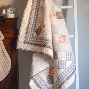 Peut inclure: Un quilt en patchwork composé de tissus crème, gris et orange doux. Le motif comprend des carrés à carreaux vichy gris et blancs, des imprimés floraux et des sections crème unies, le tout fini avec une texture de matelassage pointillée.