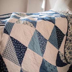 May include: A patchwork quilt featuring a repeating pattern of triangles and squares in various shades of blue, white, and navy. The fabric pieces include solid colours, small floral prints, and checkered patterns, all stitched together with visible quilting.