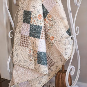May include: A patchwork quilt featuring a variety of square fabric blocks in cream, dark green, and brown plaid patterns, accented with floral prints and intricate quilted stitching, draped over a white metal rack with a wooden pumpkin nearby.