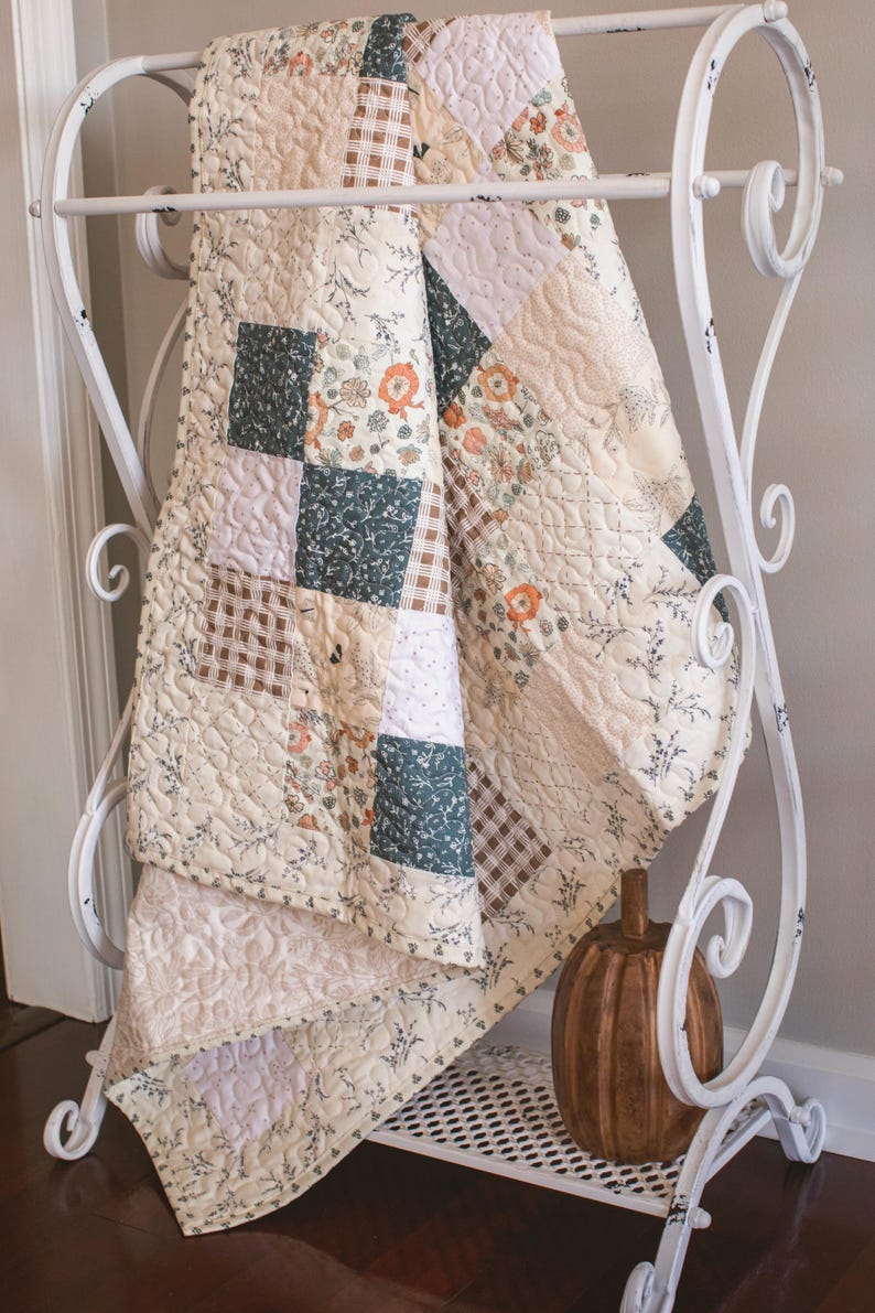 May include: A patchwork quilt featuring a variety of square fabric blocks in cream, dark green, and brown plaid patterns, accented with floral prints and intricate quilted stitching, draped over a white metal rack with a wooden pumpkin nearby.