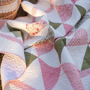 May include: A handmade quilt with a patchwork design. The quilt features white fabric with pink, green, and floral patterned accents. The edges are trimmed with a red and white gingham pattern. The quilt is folded and draped on a grassy surface.