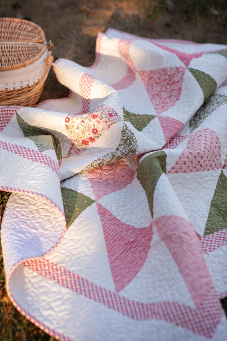 May include: A handmade quilt with a patchwork design. The quilt features white fabric with pink, green, and floral patterned accents. The edges are trimmed with a red and white gingham pattern. The quilt is folded and draped on a grassy surface.