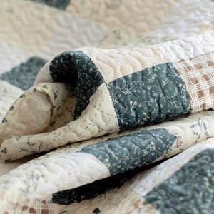 May include: A patchwork quilt featuring a variety of fabric squares in shades of dark teal, cream, and tan. The quilt displays a mix of floral patterns, solid cream textures, and plaid designs, all held together by intricate, wavy machine quilting stitches.