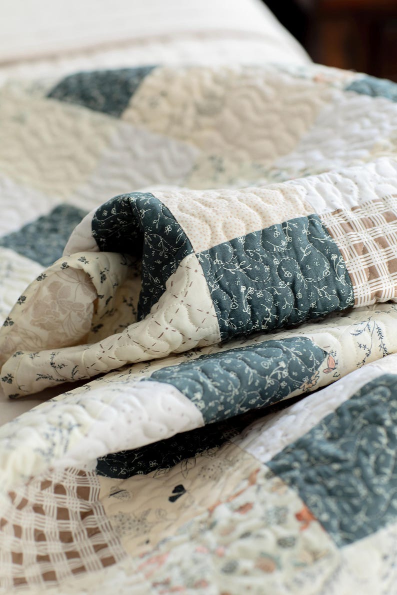 May include: A patchwork quilt featuring a variety of fabric squares in shades of dark teal, cream, and tan. The quilt displays a mix of floral patterns, solid cream textures, and plaid designs, all held together by intricate, wavy machine quilting stitches.