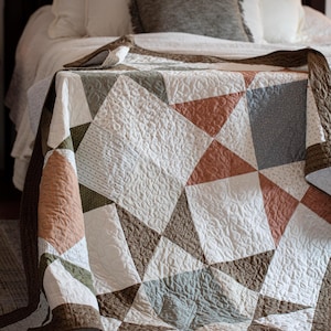 Peut inclure: Un quilt en patchwork présentant un motif d'étoile géométrique composé de blocs de tissu triangulaires et carrés dans des tons sourds de terre cuite, vert sauge, bleu ardoise et blanc, avec une texture matelassée tourbillonnante.