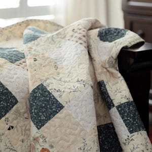 May include: A patchwork quilt featuring a repeating square pattern in shades of cream, dark blue, and soft orange. The fabric squares display various floral, botanical, and plaid prints, all unified by a consistent swirling quilted stitch pattern.