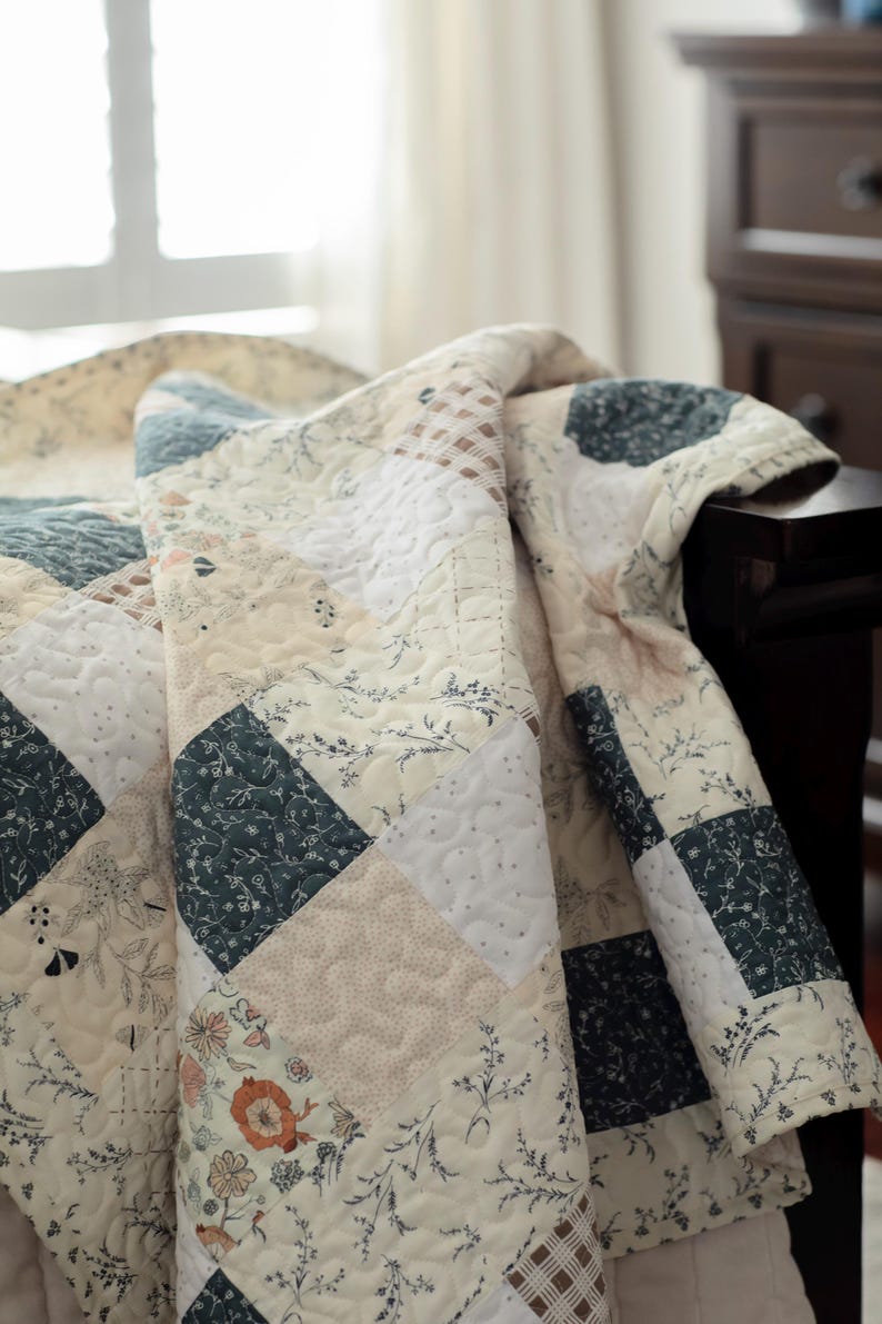 May include: A patchwork quilt featuring a repeating square pattern in shades of cream, dark blue, and soft orange. The fabric squares display various floral, botanical, and plaid prints, all unified by a consistent swirling quilted stitch pattern.