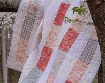May include: A patchwork quilt featuring rectangular panels in shades of coral, sage green, white, and grey. The fabric includes solid colors, floral patterns, and a grey gingham check. The quilt has white stitching in a swirling leaf pattern.