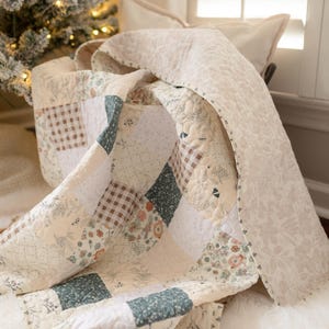 May include: A patchwork quilt featuring a variety of square fabric blocks in shades of sage green, cream, and brown. The design includes floral patterns, small polka dots, and a brown gingham check, all finished with a quilted texture and floral border.
