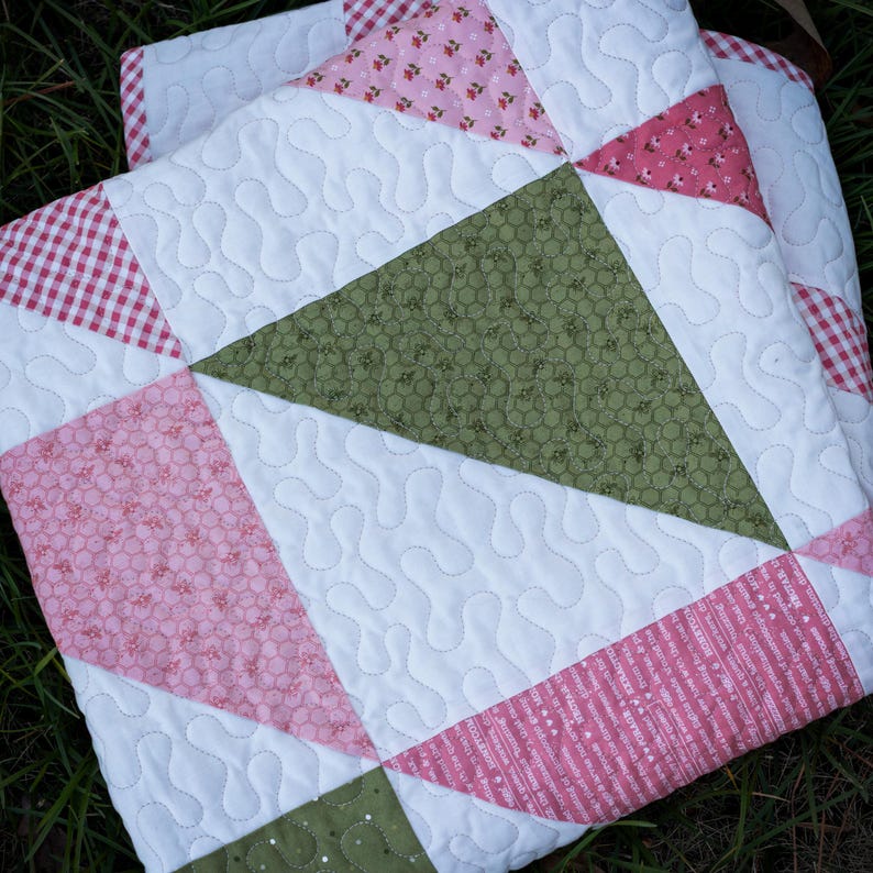 May include: A patchwork quilt in shades of white, pink, and green. The quilt has a geometric design with a wavy stitch pattern. Pink sections feature floral and striped patterns, while green sections have a dotted pattern. A cozy, handmade item.