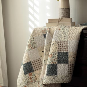 May include: A patchwork quilt featuring a variety of square fabric blocks in cream, dark green, and brown tones. The quilt displays a mix of floral patterns, small-scale geometric prints, and solid colors, all finished with a continuous swirling stitch.