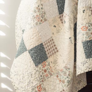May include: A patchwork quilt featuring a variety of square fabric panels in shades of cream, sage green, and muted orange. The quilt displays floral patterns, botanical illustrations, and plaid designs, all finished with a quilted texture.