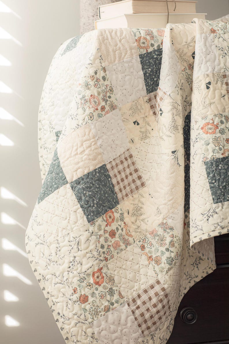 May include: A patchwork quilt featuring a variety of square fabric panels in shades of cream, sage green, and muted orange. The quilt displays floral patterns, botanical illustrations, and plaid designs, all finished with a quilted texture.