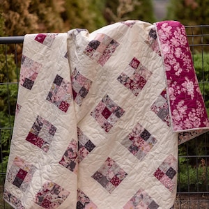 May include: A quilt featuring a cream-colored background with a repeating pattern of square blocks. Each block consists of smaller squares in shades of burgundy, pink, and grey with floral prints. The quilt has a dark pink floral border.