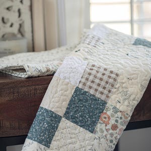 May include: A patchwork quilt featuring a variety of square fabric blocks in muted tones of cream, sage green, and brown. The quilt displays a mix of floral patterns, solid textures, and a brown plaid design, all finished with a wavy quilted stitch.