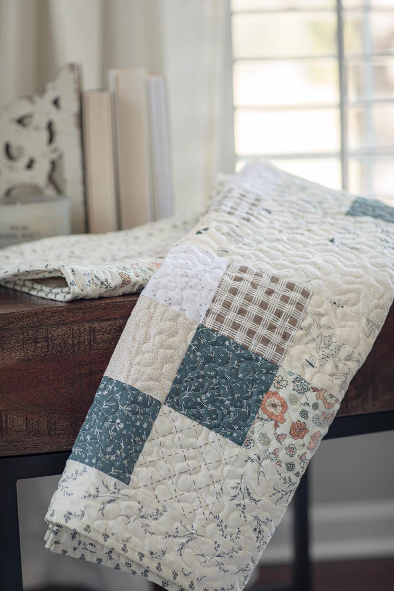 May include: A patchwork quilt featuring a variety of square fabric blocks in muted tones of cream, sage green, and brown. The quilt displays a mix of floral patterns, solid textures, and a brown plaid design, all finished with a wavy quilted stitch.
