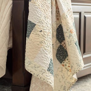 May include: A patchwork quilt featuring a variety of square fabric blocks in cream, sage green, and muted orange tones. The quilt displays floral, botanical, and plaid patterns with a textured, quilted stitch finish draped over a dark wood bed frame.