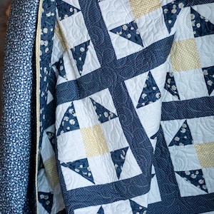 May include: A patchwork quilt featuring a repeating block pattern in navy blue, white, and soft yellow. The design incorporates floral prints, small polka dots, and a yellow gingham check, all finished with intricate swirling machine quilting stitches.