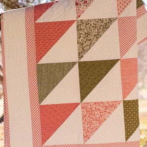 May include: A patchwork quilt with a variety of patterns and colours. The quilt features coral, olive green, and floral patterned triangles, set against a white background. The border is a red and white gingham pattern.