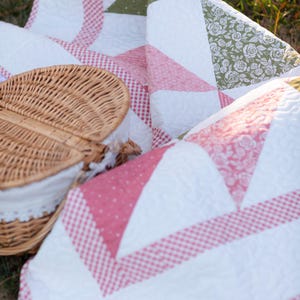 May include: A picnic scene featuring a white and pink quilt with a gingham pattern and floral accents. A woven picnic basket with a white lace-trimmed lining is partially visible, suggesting a relaxing outdoor setting.