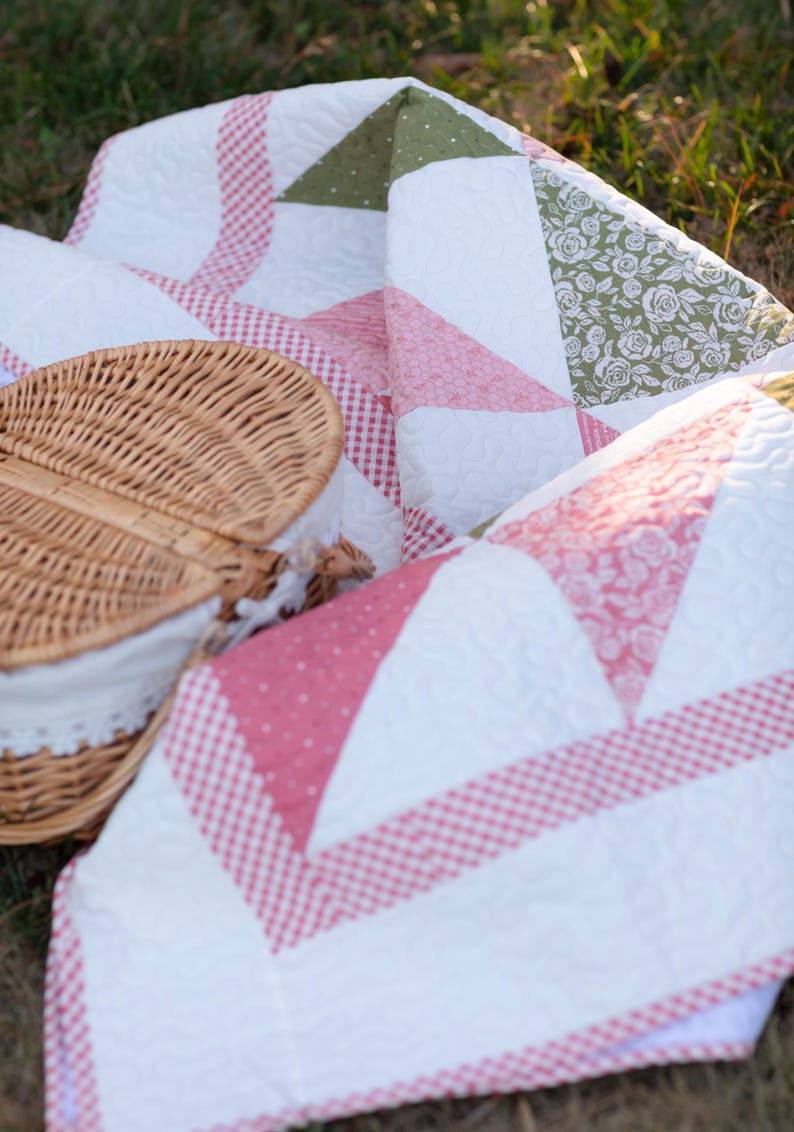 May include: A picnic scene featuring a white and pink quilt with a gingham pattern and floral accents. A woven picnic basket with a white lace-trimmed lining is partially visible, suggesting a relaxing outdoor setting.