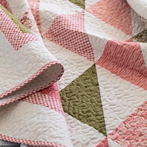 May include: A close-up of a handmade quilt with a patchwork design. The quilt features white, pink, and green triangles, with a red and white gingham border. The quilt is folded, showing the detailed stitching and textures.