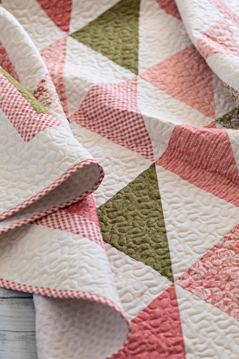 May include: A close-up of a handmade quilt with a patchwork design. The quilt features white, pink, and green triangles, with a red and white gingham border. The quilt is folded, showing the detailed stitching and textures.