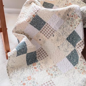 May include: A patchwork quilt featuring a variety of square fabric blocks in muted tones of sage green, cream, and brown. The design includes floral patterns, small-scale gingham checks, and solid cream squares, all finished with a quilted texture.