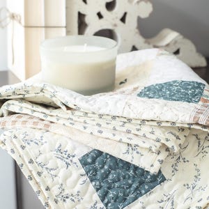 May include: A folded patchwork quilt featuring a cream-colored base with dark teal floral patterns, light brown checkered squares, and intricate white stitching. The fabric displays a repeating botanical print with delicate stems and leaves.