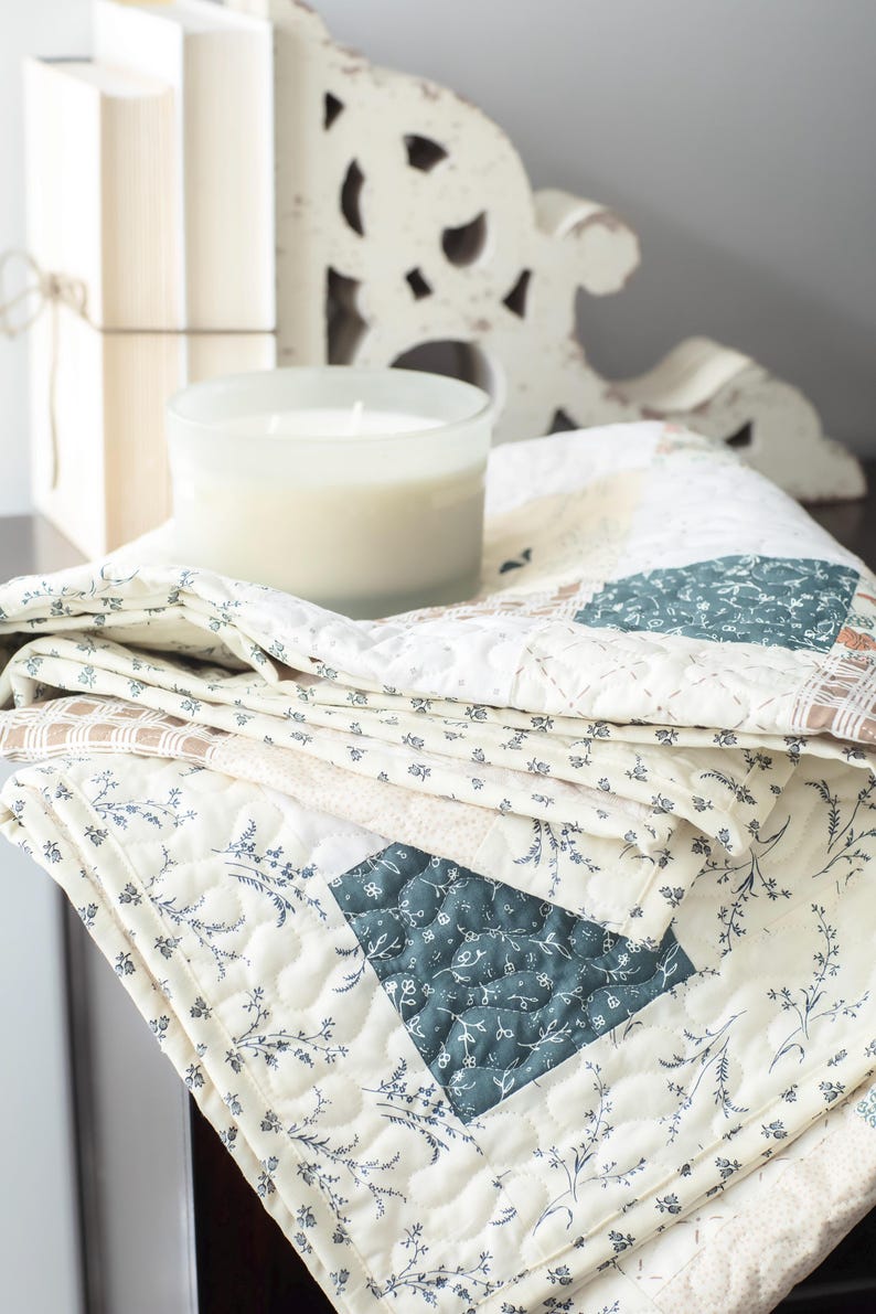 May include: A folded patchwork quilt featuring a cream-colored base with dark teal floral patterns, light brown checkered squares, and intricate white stitching. The fabric displays a repeating botanical print with delicate stems and leaves.