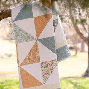 Peut inclure: Une courtepointe faite à la main avec un motif géométrique de triangles blancs, turquoise et pêche, ornée d'accents floraux et à pois. La courtepointe est drapée sur une branche d'arbre, mettant en valeur son design et sa texture.