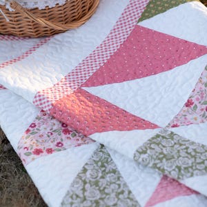 May include: A white quilt with pink, green, and floral patterned triangles. The quilt has a red and white gingham border and a basket with a lid. The quilt is likely for a picnic or outdoor use.