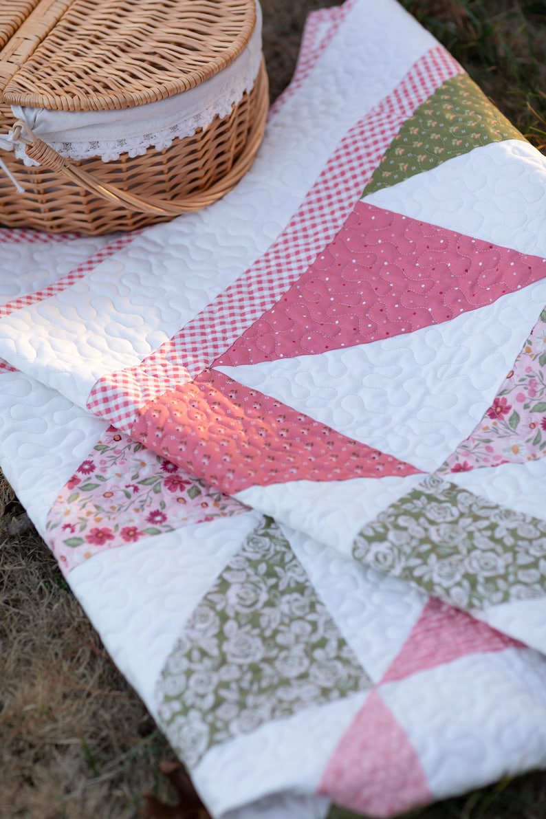May include: A white quilt with pink, green, and floral patterned triangles. The quilt has a red and white gingham border and a basket with a lid. The quilt is likely for a picnic or outdoor use.