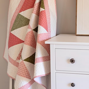 May include: A handmade quilt with a geometric design, featuring triangles in white, pink, and olive green. The quilt is draped over a white ladder. The edges are trimmed with a red and white checkered pattern. The quilt is next to a white chest of drawers.