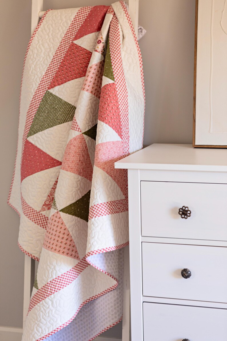 May include: A handmade quilt with a geometric design, featuring triangles in white, pink, and olive green. The quilt is draped over a white ladder. The edges are trimmed with a red and white checkered pattern. The quilt is next to a white chest of drawers.