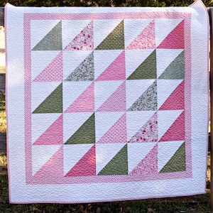 May include: A handmade quilt with a geometric design. The quilt features alternating triangles in shades of pink and green, set against a white background. A pink dotted border frames the quilt, which is likely made of cotton.